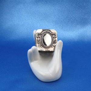 May include: A small, square, silver-toned metal box featuring an embossed oval center panel surrounded by intricate floral and geometric border patterns, held within a white ceramic hand-shaped display stand for jewelry or small trinkets.
