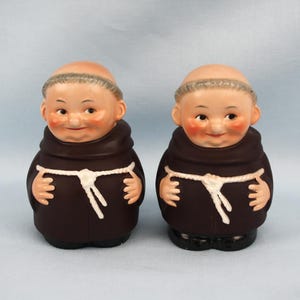 Puede incluir: Dos figuras de cerámica de monjes. Cada figura tiene la cabeza calva, mejillas rosadas y una túnica marrón con un cinturón de cuerda blanco. Las figuras están una al lado de la otra, sobre un fondo azul claro.