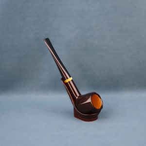 Puede incluir: Una pipa de tabaco marrón oscuro con una banda amarilla cerca de la boquilla. La pipa tiene una caña curvada y un cuenco redondeado, sobre un fondo azul grisáceo. El interior del cuenco es de un marrón más claro.
