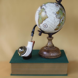 May include: A vintage globe with a detailed map of the world sits on a wooden stand, next to a white ceramic pipe with a brass lid. Both items are displayed on a green book with a brown cover.