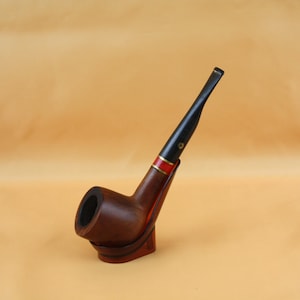 May include: A wooden tobacco pipe with a dark brown bowl and stem. The pipe has a black mouthpiece and a red and gold band. The pipe is resting on a matching wooden stand.