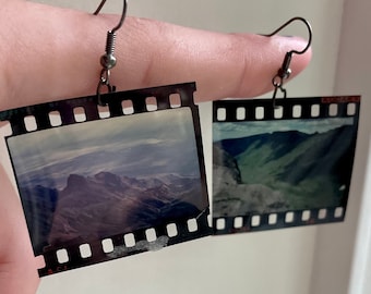 Puede incluir: Un par de pendientes colgantes formados por segmentos rectangulares de tiras de película de 35mm vintage. Cada pendiente muestra una fotografía de paisaje con montañas y valles, enmarcada por bordes negros con perforaciones y el texto KODAK.