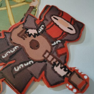May include: A stack of die-cut stickers featuring cartoon characters. One sticker depicts a figure in brown and red attire playing a ukulele. The other sticker shows a figure in green attire holding a torch. The stickers have a red border.