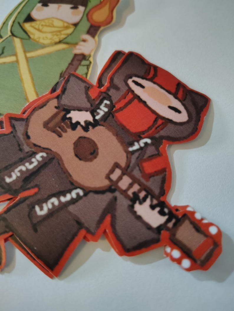 May include: A stack of die-cut stickers featuring cartoon characters. One sticker depicts a figure in brown and red attire playing a ukulele. The other sticker shows a figure in green attire holding a torch. The stickers have a red border.