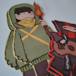 May include: Two paper cutouts of cartoon figures. One figure is wearing a green hooded sweatshirt, a yellow scarf, and holding a torch with a flame. The other figure is wearing a red and black outfit and holding a guitar. The figures are on a white background.