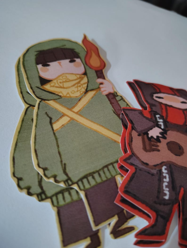 May include: Two paper cutouts of cartoon figures. One figure is wearing a green hooded sweatshirt, a yellow scarf, and holding a torch with a flame. The other figure is wearing a red and black outfit and holding a guitar. The figures are on a white background.