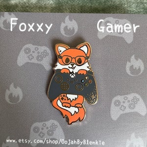 May include: A pin featuring a cartoon fox wearing glasses and holding a gray video game controller. The pin is on a gray card with the words "Foxxy Gamer" and website addresses. The fox is orange, white, and black.