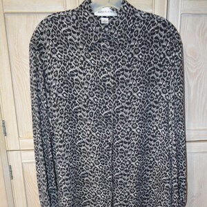 May include: A long-sleeved, black and white leopard print blouse with a button-up front and a collar. The blouse is made of a lightweight fabric and has a relaxed fit. The label on the blouse reads "Anne Klein".