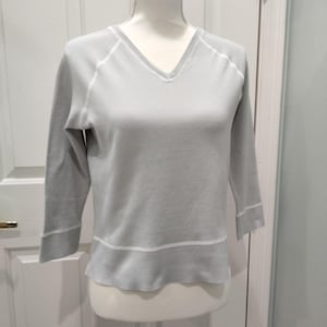 May include: A light gray V-neck sweater with three-quarter sleeves. The sweater features white stitching along the neckline, shoulder seams, and sleeve cuffs. A white band accents the bottom hem. The sweater is displayed on a mannequin.