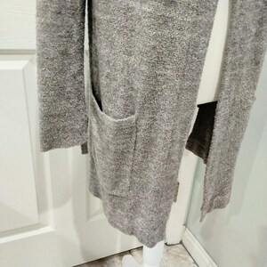 Pode incluir: Um cardigan longo e aberto em tecido cinza mesclado e macio. O su&eacute;ter apresenta dois bolsos frontais e mangas compridas. A pe&ccedil;a &eacute; exibida em um manequim branco, mostrando sua queda e textura.