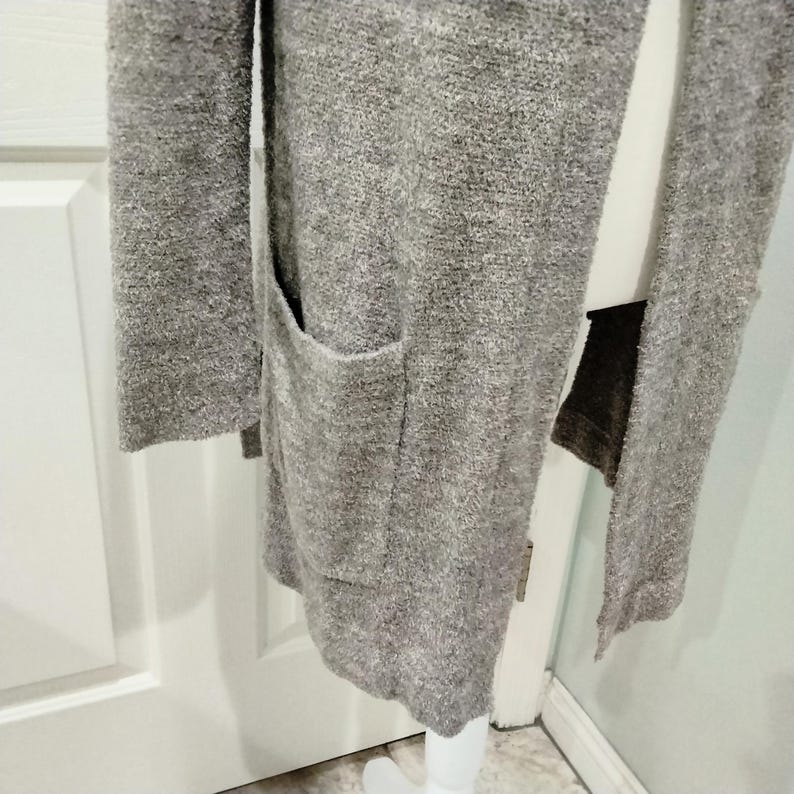 Pode incluir: Um cardigan longo e aberto em tecido cinza mesclado e macio. O su&eacute;ter apresenta dois bolsos frontais e mangas compridas. A pe&ccedil;a &eacute; exibida em um manequim branco, mostrando sua queda e textura.