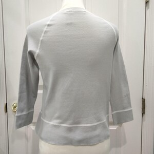May include: A light gray sweater with three-quarter sleeves. The sweater has white trim along the cuffs and hem, and white stitching along the raglan sleeves. The neckline is a simple crew neck. The sweater is displayed on a mannequin.