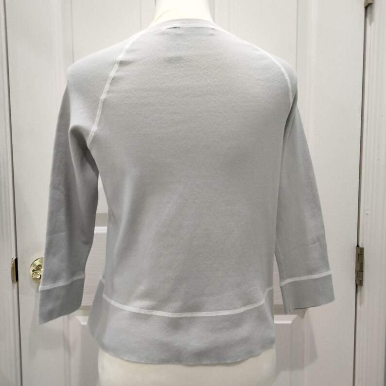 May include: A light gray sweater with three-quarter sleeves. The sweater has white trim along the cuffs and hem, and white stitching along the raglan sleeves. The neckline is a simple crew neck. The sweater is displayed on a mannequin.