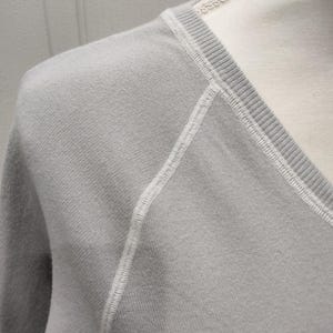 May include: Close-up of a light gray V-neck sweater. The sweater features white stitching along the seams and neckline. The fabric appears soft and has a subtle texture. The sweater is displayed on a mannequin.