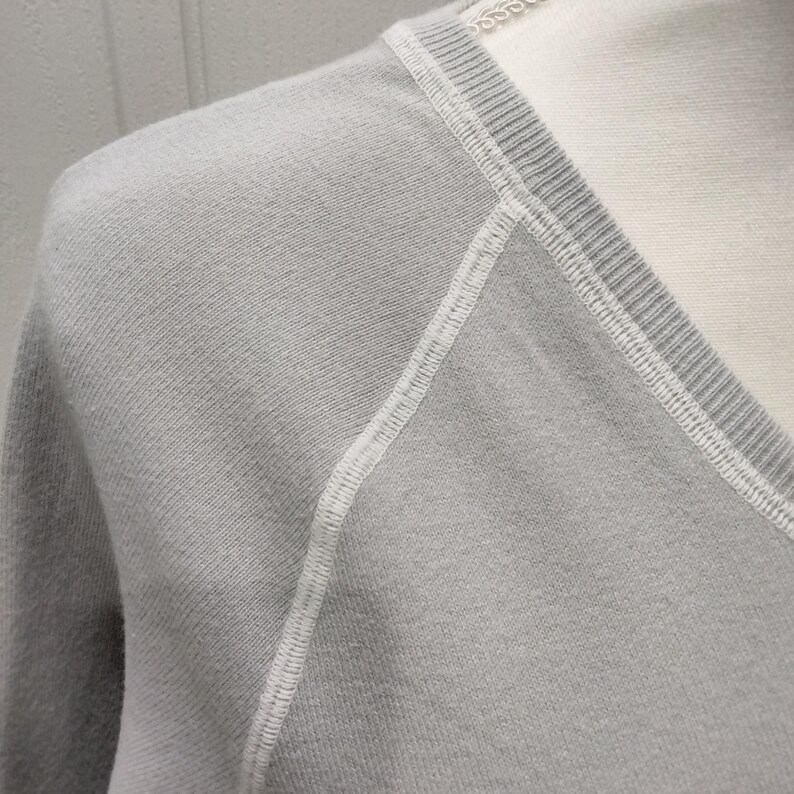 May include: Close-up of a light gray V-neck sweater. The sweater features white stitching along the seams and neckline. The fabric appears soft and has a subtle texture. The sweater is displayed on a mannequin.