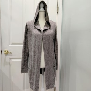 Pode incluir: Um cardigan longo e aberto na frente, em cinza mesclado, com capuz e dois bolsos frontais. O cardigan &eacute; exibido em um manequim, mostrando seu ajuste descontra&iacute;do e estilo casual. A pe&ccedil;a &eacute; feita de um material macio e texturizado.