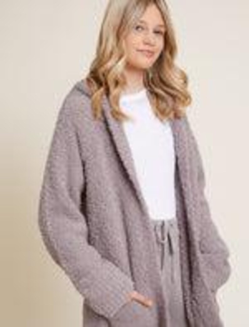 Pode incluir: Um cardigan longo e aberto em malha mauve macia e texturizada. O cardigan apresenta um capuz, dois bolsos frontais e um ajuste descontra&iacute;do. As mangas s&atilde;o longas e o tecido parece aconchegante e quente, adequado para usar em camadas.
