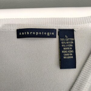 May include: Close-up of a light gray sweater with the brand tag "Anthropologie" visible. A second tag indicates the sweater is size L and made of cotton, nylon, and lycra. The tag also states the sweater was made in Hong Kong.
