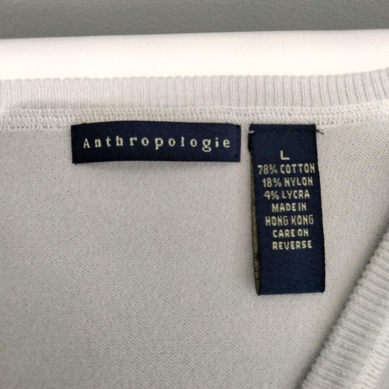 May include: Close-up of a light gray sweater with the brand tag "Anthropologie" visible. A second tag indicates the sweater is size L and made of cotton, nylon, and lycra. The tag also states the sweater was made in Hong Kong.