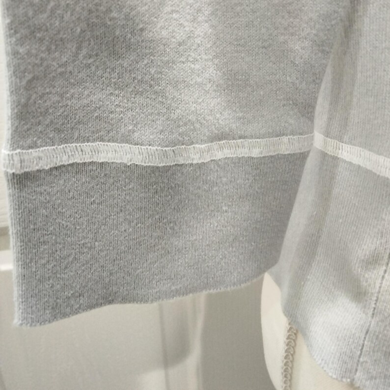 May include: Close-up of a gray sweatshirt sleeve. The cuff is a darker shade of gray and is stitched with white thread. The fabric has a ribbed texture.