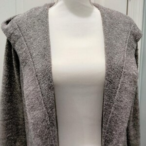 Pode incluir: Um cardigan cinza com capuz exibido em um manequim. O cardigan tem uma apar&ecirc;ncia texturizada e um design de frente aberta. O capuz &eacute; vis&iacute;vel na parte superior, e a pe&ccedil;a parece ser feita de um material macio e confort&aacute;vel.