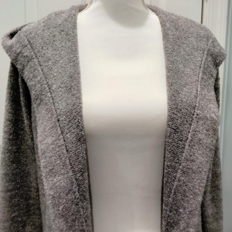 Pode incluir: Um cardigan cinza com capuz exibido em um manequim. O cardigan tem uma apar&ecirc;ncia texturizada e um design de frente aberta. O capuz &eacute; vis&iacute;vel na parte superior, e a pe&ccedil;a parece ser feita de um material macio e confort&aacute;vel.