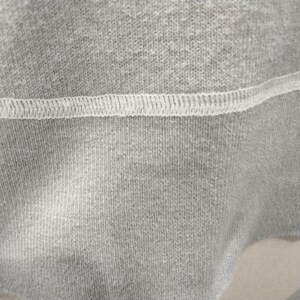 May include: Close-up of gray fabric with a white stitched seam. The fabric has a textured appearance, with visible vertical lines. The seam is a series of small, evenly spaced stitches, creating a contrasting white line across the gray material.