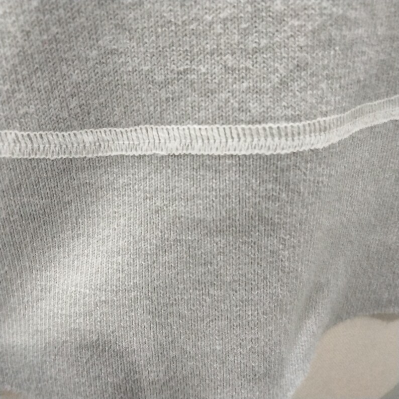 May include: Close-up of gray fabric with a white stitched seam. The fabric has a textured appearance, with visible vertical lines. The seam is a series of small, evenly spaced stitches, creating a contrasting white line across the gray material.