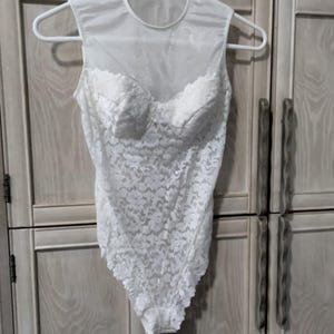 May include: White lace bodysuit with a sheer neckline and cap sleeves, hanging on a white hanger. The bodice features a floral lace pattern and a sweetheart neckline. The garment is a one-piece design.