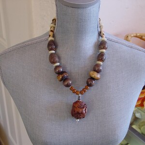 May include: A brown beaded necklace with a carved pendant. The necklace features various sizes of brown, tan, and cream-colored beads. The pendant is a carved figure in a reddish-brown hue, suspended from a silver-toned bail.