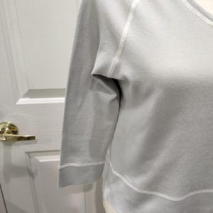 May include: A light gray sweatshirt with three-quarter sleeves. The sleeves and hem are accented with white stitching. The neckline is a scoop neck. The sweatshirt is made of a soft material.