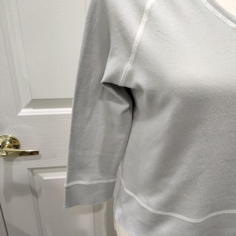 May include: A light gray sweatshirt with three-quarter sleeves. The sleeves and hem are accented with white stitching. The neckline is a scoop neck. The sweatshirt is made of a soft material.
