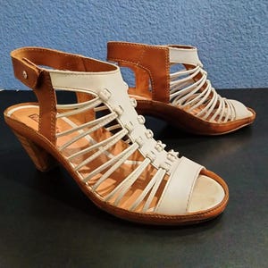 May include: A pair of open-toe sandals with a white and brown leather design. The sandals have a strappy upper, a small heel, and a buckle closure on the ankle strap. The shoes are displayed on a dark surface.