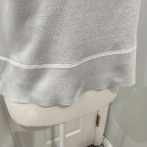 May include: Close-up of a light gray sweatshirt with a white stitched hemline. The bottom edge of the sweatshirt has a raw, unfinished look. The garment is displayed on a white mannequin.