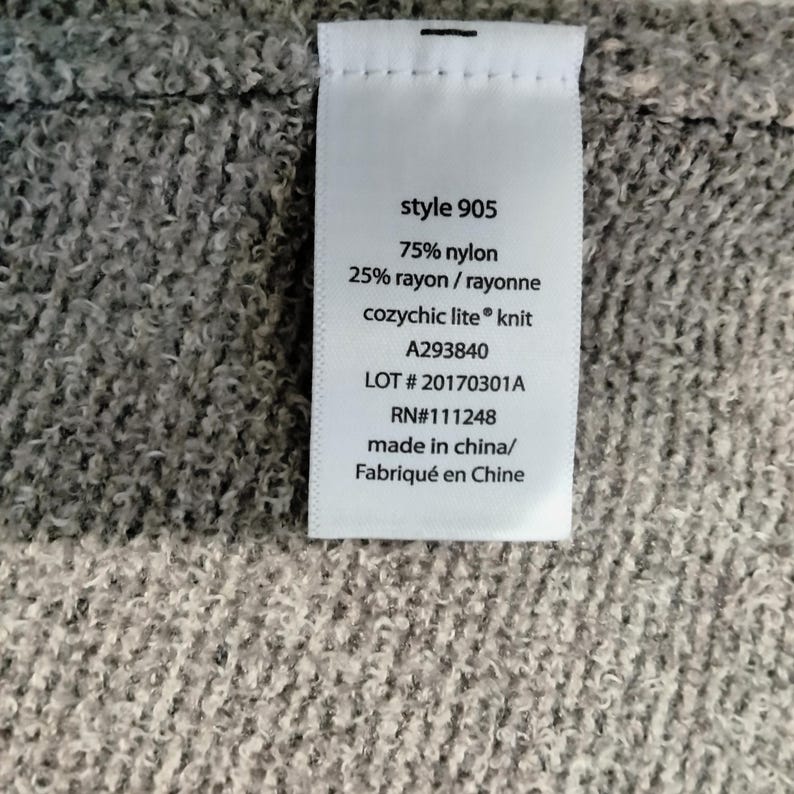 Pode incluir: Close-up de uma etiqueta de roupa branca com texto preto. A etiqueta diz "style 905", lista a composi&ccedil;&atilde;o do tecido como 75% nylon e 25% rayon, e inclui a marca "cozychic lite knit". Detalhes adicionais incluem um n&uacute;mero de lote e RN.
