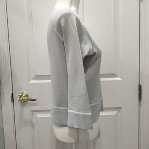 May include: A light gray sweater with long sleeves and a crew neck. The sweater has white trim at the cuffs and hem. The garment is displayed on a mannequin against a white door.
