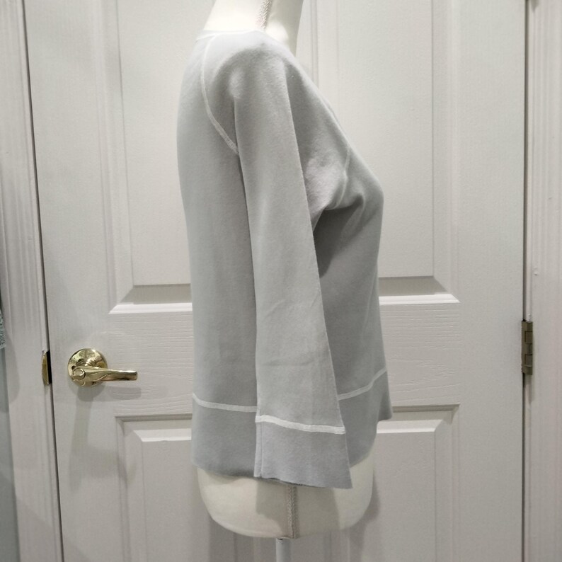 May include: A light gray sweater with long sleeves and a crew neck. The sweater has white trim at the cuffs and hem. The garment is displayed on a mannequin against a white door.