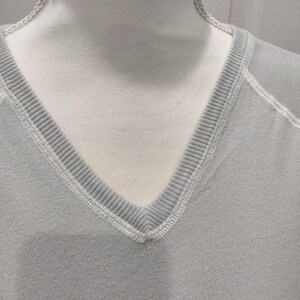 May include: Close-up of a light gray V-neck sweatshirt. The neckline is ribbed with white stitching. The fabric appears to be a soft knit material. The sweatshirt is displayed on a mannequin.