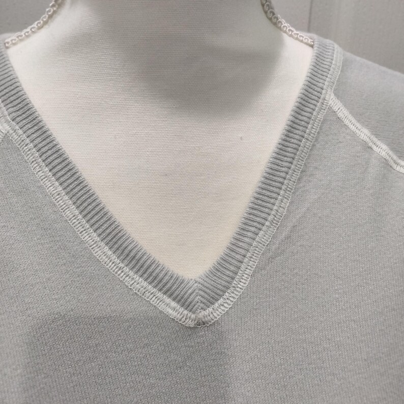 May include: Close-up of a light gray V-neck sweatshirt. The neckline is ribbed with white stitching. The fabric appears to be a soft knit material. The sweatshirt is displayed on a mannequin.