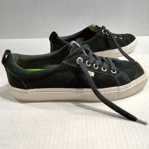 May include: A pair of black suede sneakers with white soles and white laces. The shoes have a low-profile design with a rounded toe and a textured sole. The laces are threaded through silver eyelets. The sneakers are casual footwear.