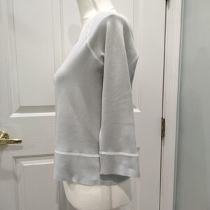 May include: A light gray long-sleeve shirt with a V-neck and white trim. The shirt has a relaxed fit and three-quarter sleeves. The hem and neckline are accented with a thin white band.