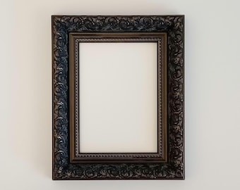 Puede incluir: Un marco de imagen de color marrón oscuro adornado con un paspartú blanco y un espacio blanco vacío en el interior. El marco tiene un diseño decorativo e intrincado.