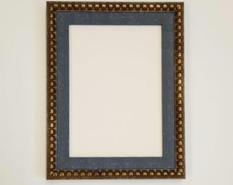 May include: Rectangular picture frame with a gold-colored beaded border and a dark blue inner frame. The frame holds a white space for a photo or artwork. The frame is displayed against a white wall.