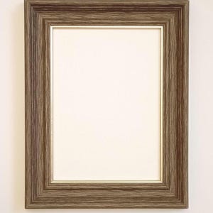 May include: A rectangular picture frame with a brown wood grain finish. The frame has a wide, beveled edge and a thin, gold-coloured inner border. The centre is a cream-coloured matting, ready for a photo or artwork.