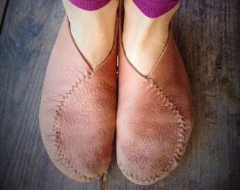 May include: A pair of light brown leather slippers with visible stitching details. The slippers are worn by a person wearing purple leggings. The slippers are on a wooden surface.