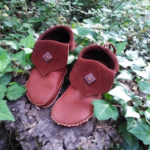 May include: A pair of brown leather moccasin-style baby shoes with a folded-over cuff. The cuffs are a darker shade of brown and feature a small, square decorative element. The shoes are set on a weathered wooden surface, surrounded by green foliage.