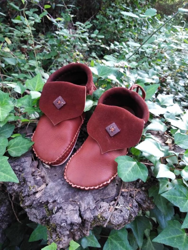 May include: A pair of brown leather moccasin-style baby shoes with a folded-over cuff. The cuffs are a darker shade of brown and feature a small, square decorative element. The shoes are set on a weathered wooden surface, surrounded by green foliage.