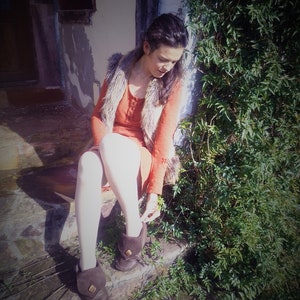 May include: A person wearing a rust-colored dress and a faux fur vest sits on stone steps. They are adjusting the strap of a pair of brown leather boots. Green foliage is visible on the right side of the image.