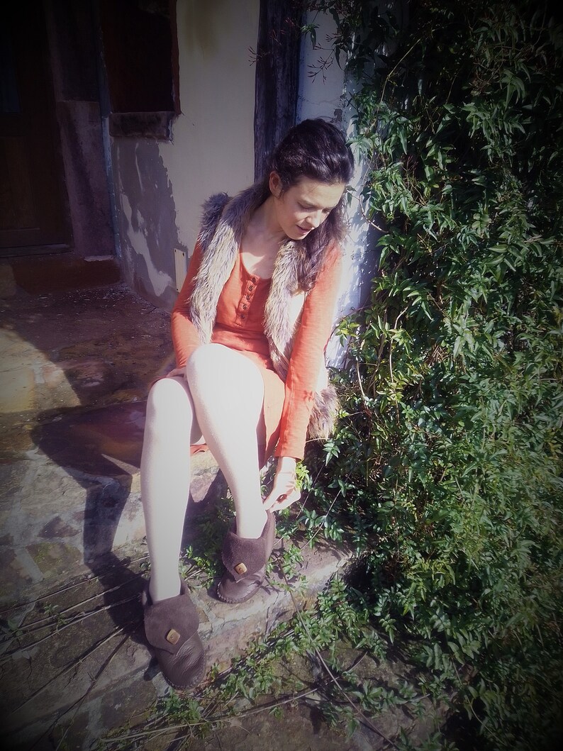 May include: A person wearing a rust-colored dress and a faux fur vest sits on stone steps. They are adjusting the strap of a pair of brown leather boots. Green foliage is visible on the right side of the image.