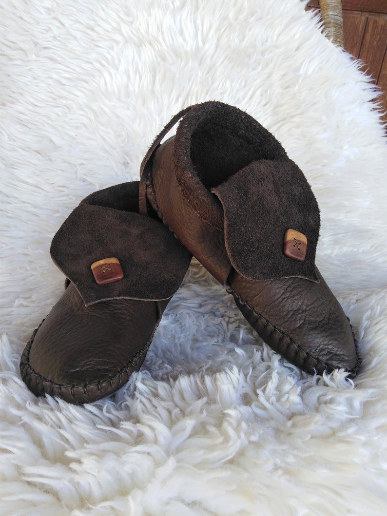 May include: A pair of brown leather moccasin-style shoes with a folded-over flap secured by a wooden button. The shoes have a stitched sole and a strap at the back. They are displayed on a white, fluffy surface.